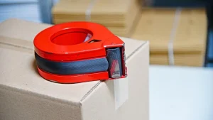 Red tape dispenser on a sealed cardboard box, with packed shipping cartons stacked behind it